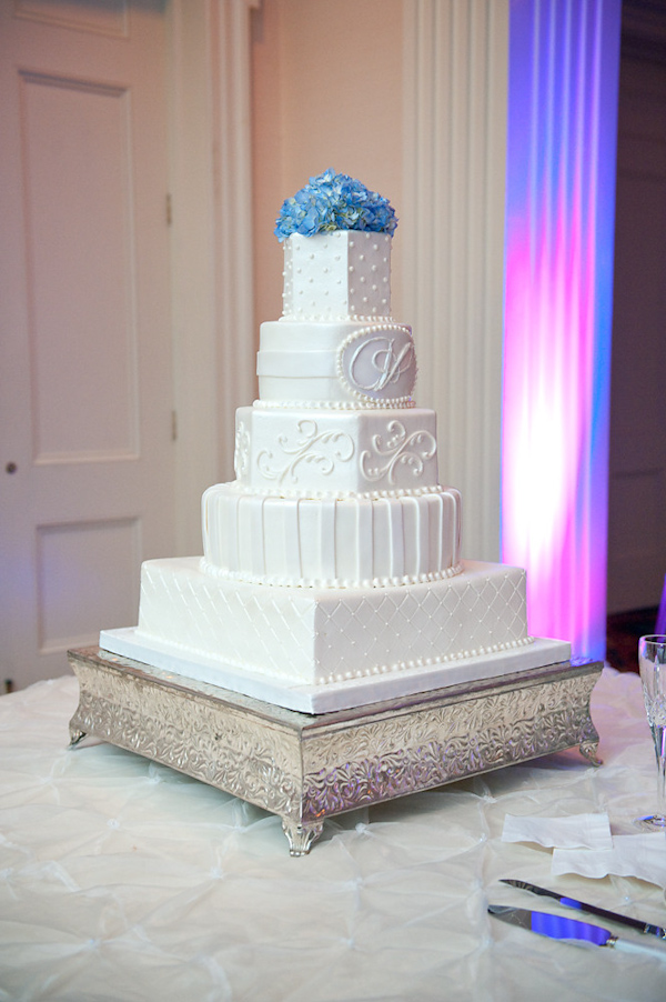white four tiered wedding cake on vintage style cake stand with different patterned layers, monogram design, and blue floral cake topper with purple and blue lighting - photo by Houston based wedding photographer Adam Nyholt